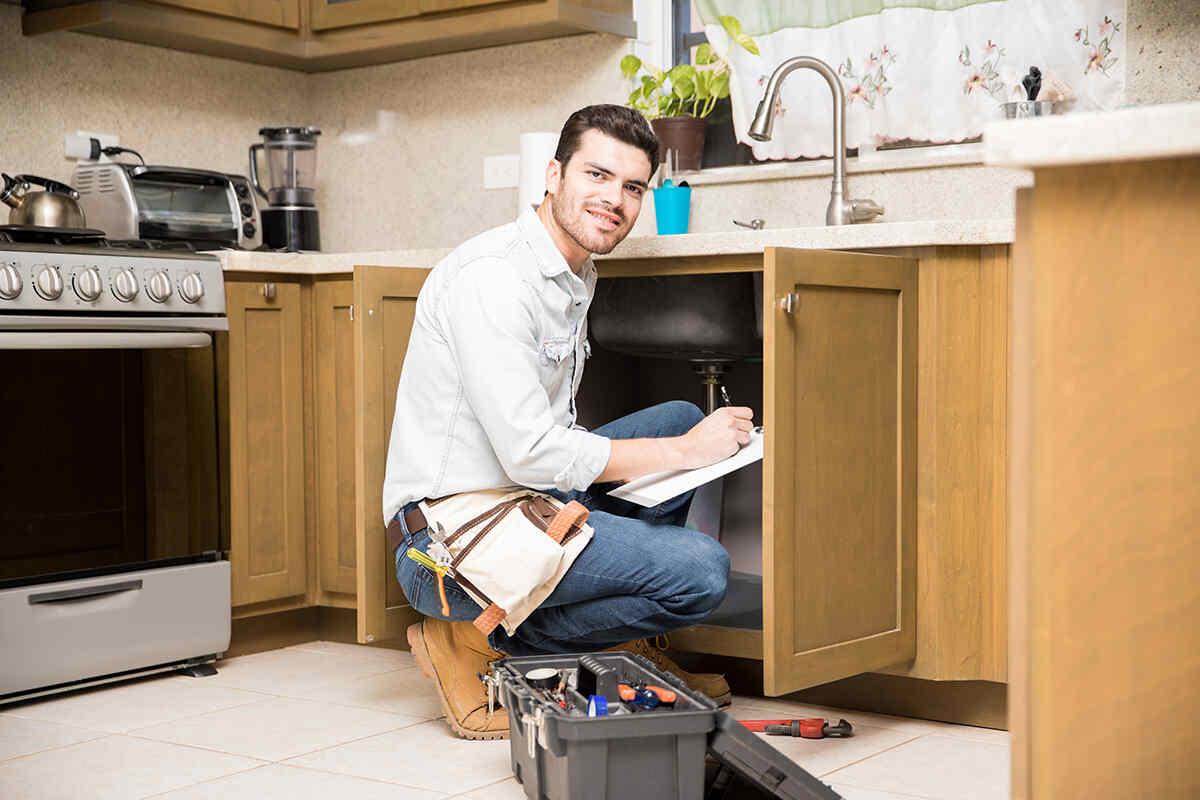 A plumber kneeling by an open kitchen cabinet writing an inspection report on a clipboard.